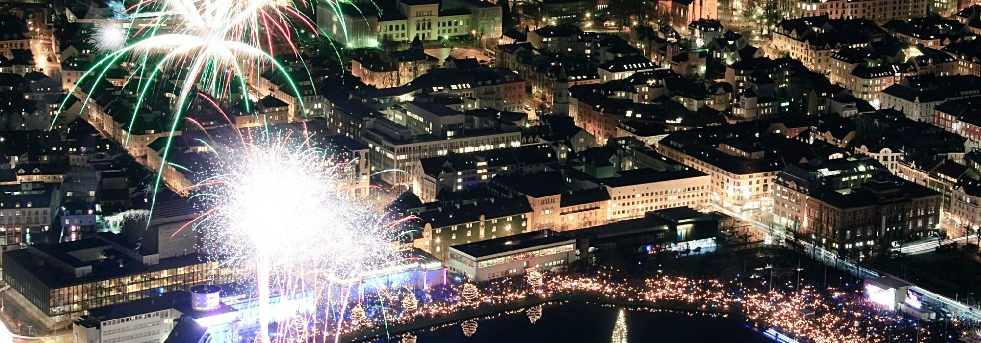 Luftfoto av en by om natten med fyrverkeri som lyser opp himmelen over bygninger og en stor opplyst innsjø omgitt av lys.