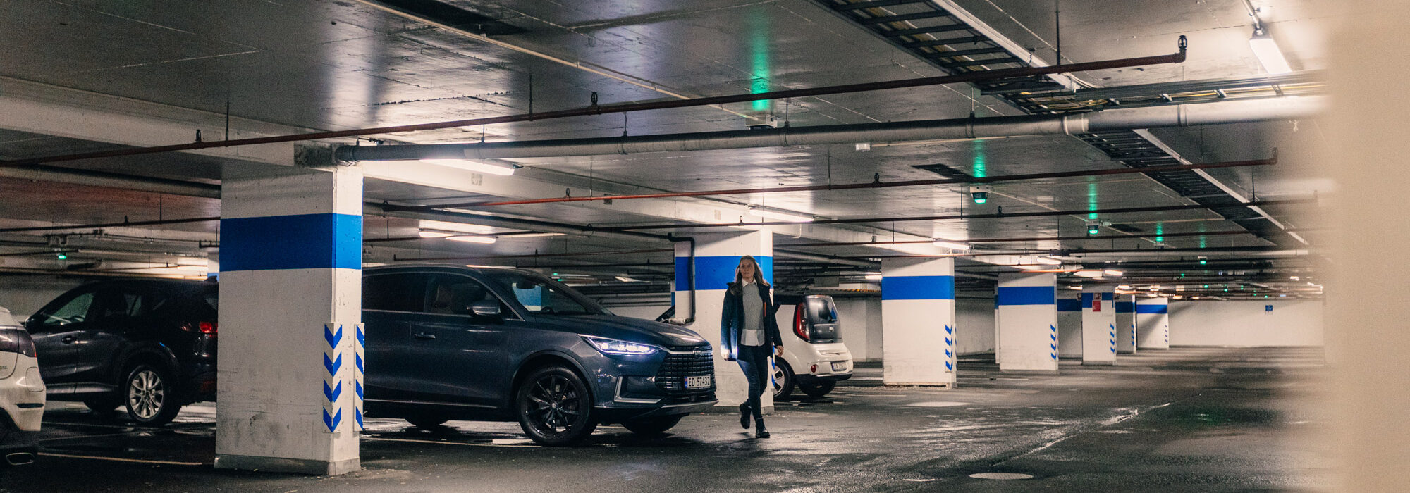 Bergen-Parkering-209 En person som går i et underjordisk parkeringshus med biler parkert på begge sider og overlys som lyser opp området.