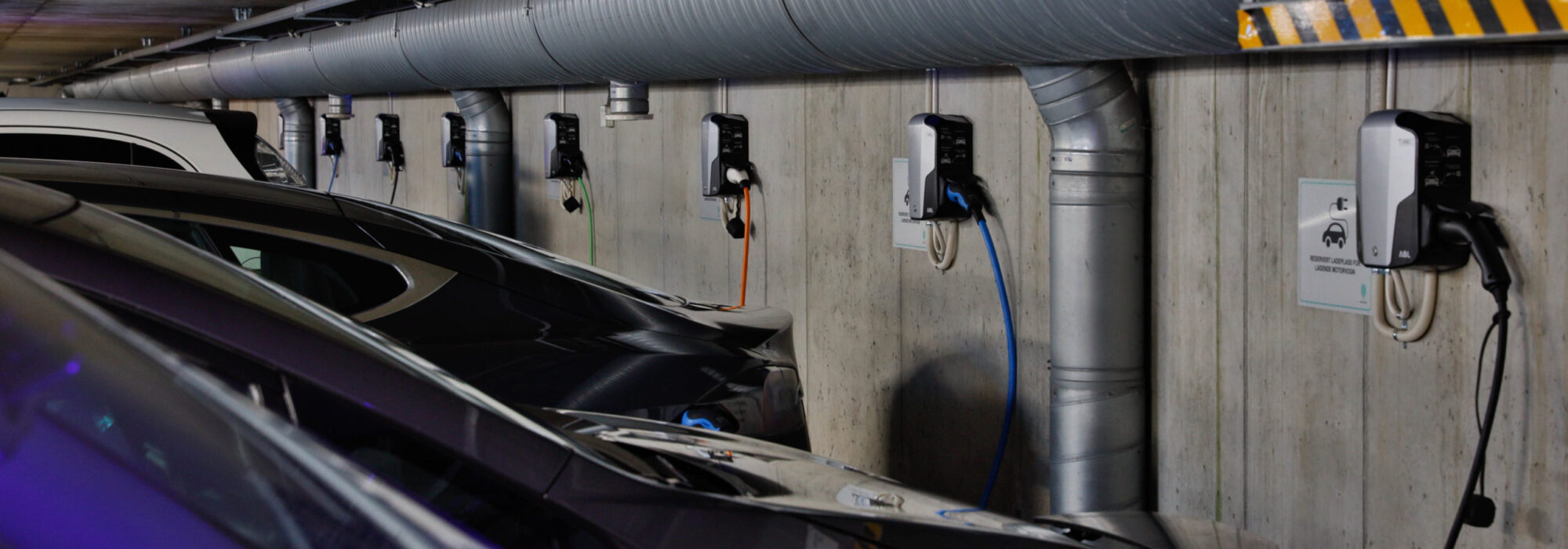 Bergen-Parkering-Foto-G-Fotobloggo-AHR-28 Biler parkert i en garasje, hver koblet til en veggmontert ladestasjon for elektriske kjøretøy.