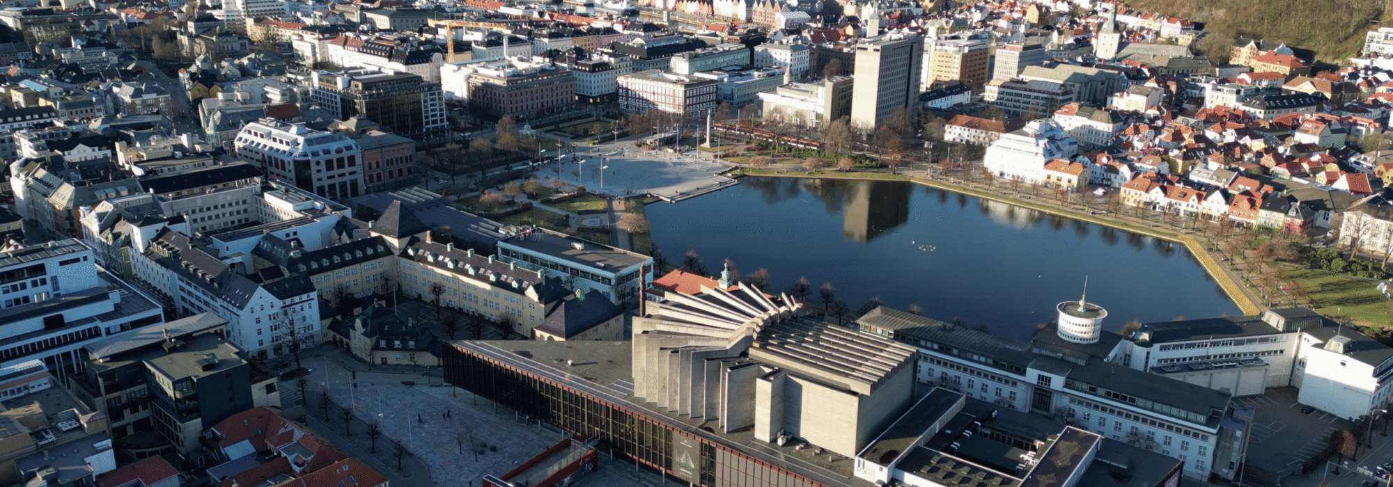 Flyfoto av sentrum i Bergen, Norge, som viser byens bygninger, Lille Lungegårdsvann, Vågen og de omkringliggende byfjellene under klar himmel.