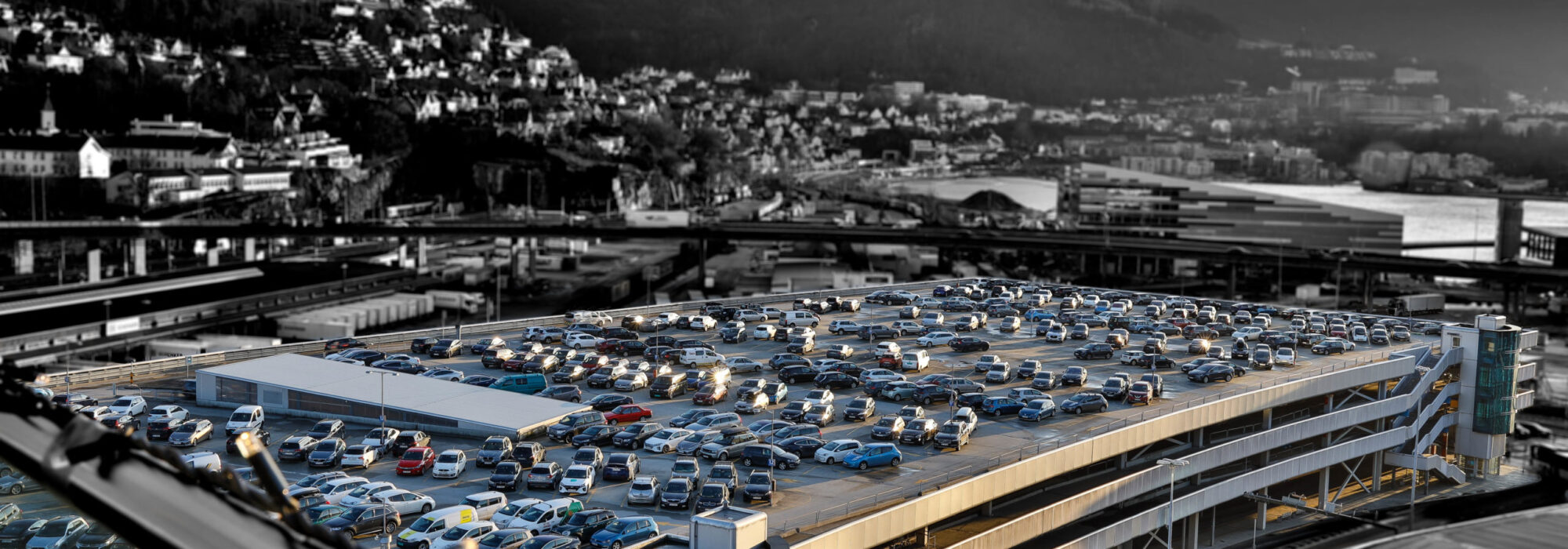 Et flerplans parkeringshus fylt med biler ligger i Bergen sentrum, et urbant område med bakker i bakgrunnen under en delvis overskyet himmel.