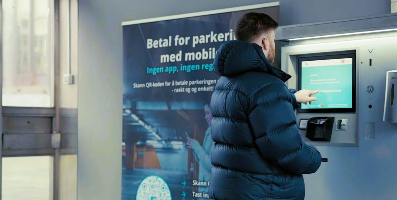 En mann i mørk pufferjakke bruker en parkeringsautomat med berøringsskjerm ved siden av et skilt med instruksjoner på norsk.