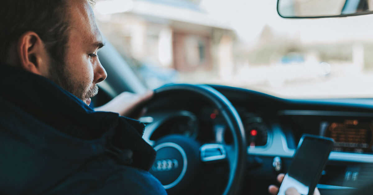 bilogmotormagasinet En sjåfør ser på en smarttelefon mens han tar tak i rattet, med et uskarpt Bergen sentrum synlig gjennom frontruten. Husk, ikke stress og hold fokus på veien videre for å sikre alles sikkerhet.