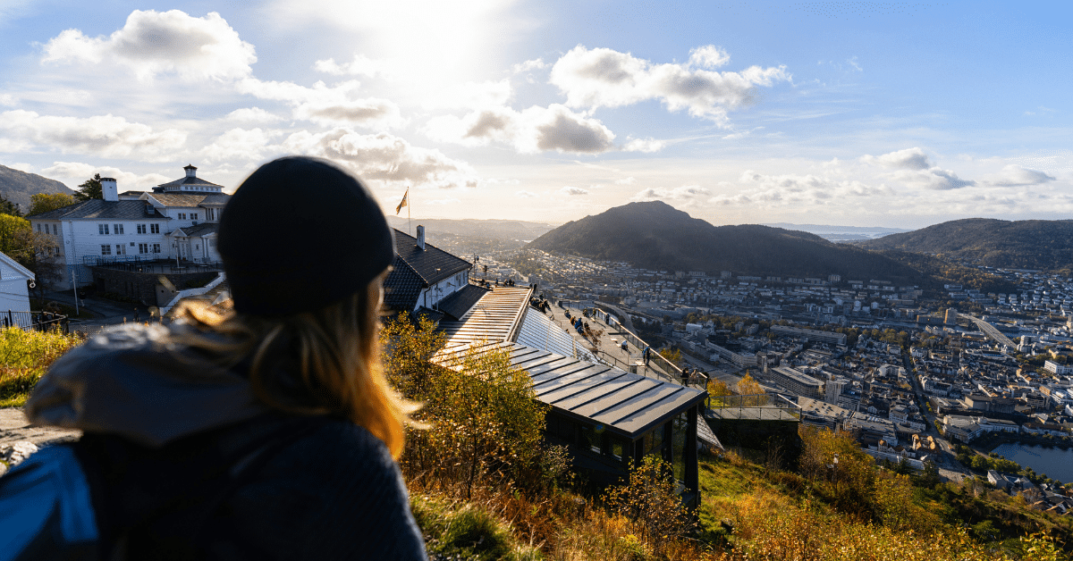 En person med ryggsekk og lue ser ut over et bylandskapet og byfjellene fra Fløyen, under en lys, delvis overskyet himmel.