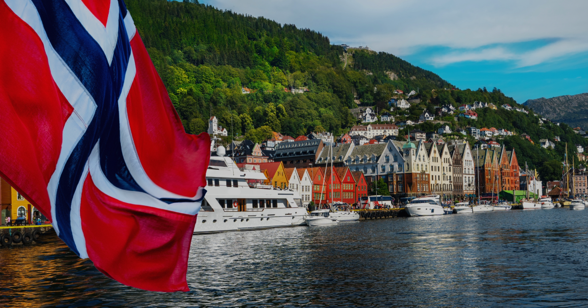 feat-img-17-mai-2024 Norsk flagg i forgrunnen med havn, båter, tradisjonelle trebygninger og skogkledde åser i bakgrunnen under en delvis overskyet himmel.