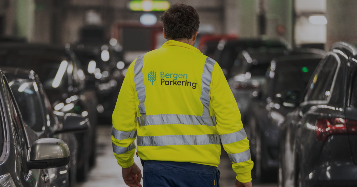 feat-img-ISO14001 Person i synlig jakke med "Bergen Parkering" på baksiden går gjennom et svakt opplyst parkeringshus omgitt av biler.