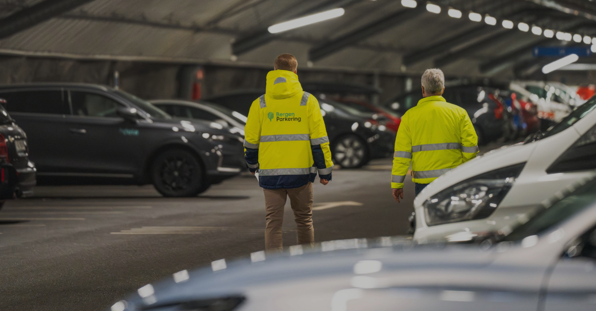 feat-img-apenhetsloven-2024-1 To parkeringsvakter i synlige jakker går gjennom et overbygd parkeringshus fylt med parkerte biler.