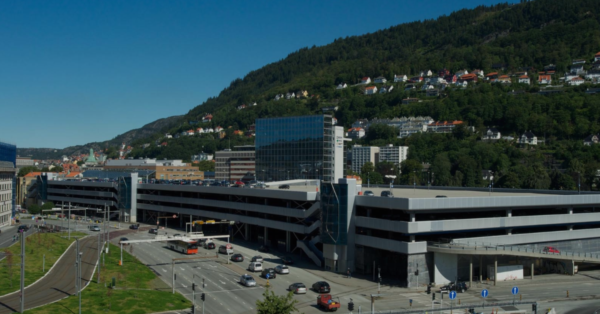 En bygning på flere plan med en parkeringsstruktur foran, omgitt av en vei med biler. Grønne åser og spredte hus er i bakgrunnen under en klar blå himmel.