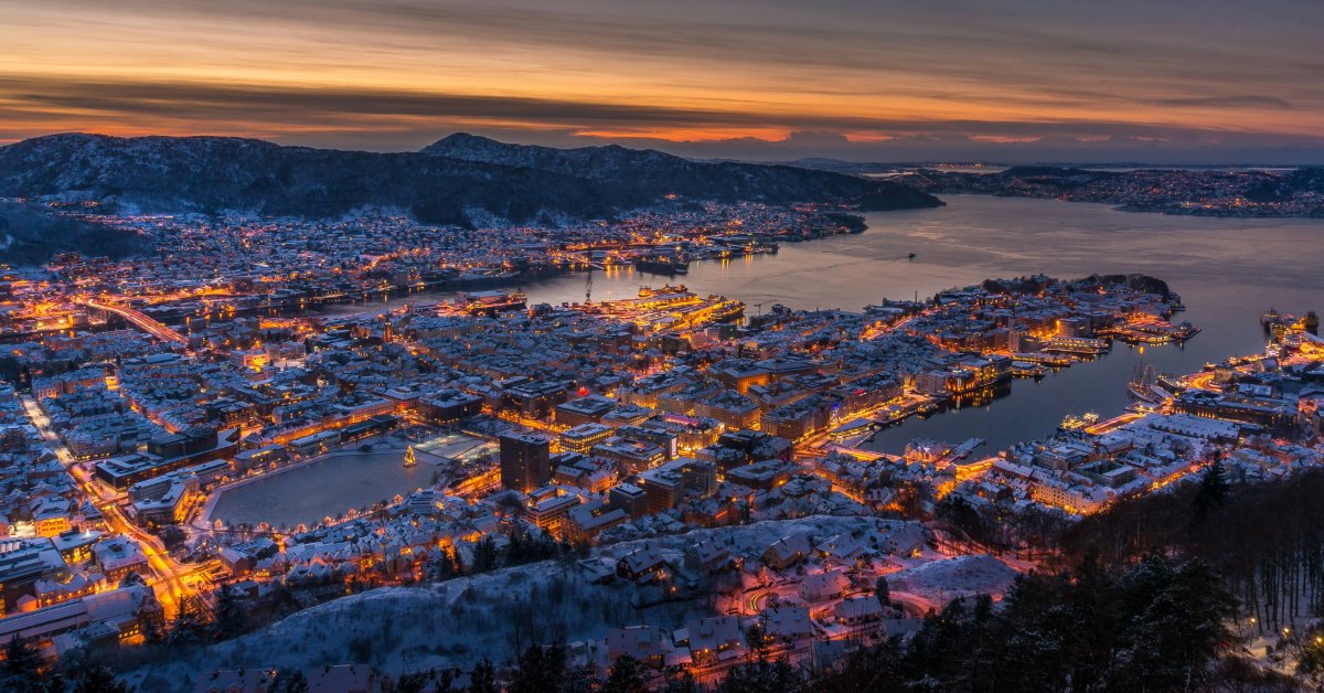 Luftfoto av en snødekt by i skumringen, omgitt av vann og åser. Byen er opplyst med gatelys, og himmelen er en gradient av oransje og lilla.