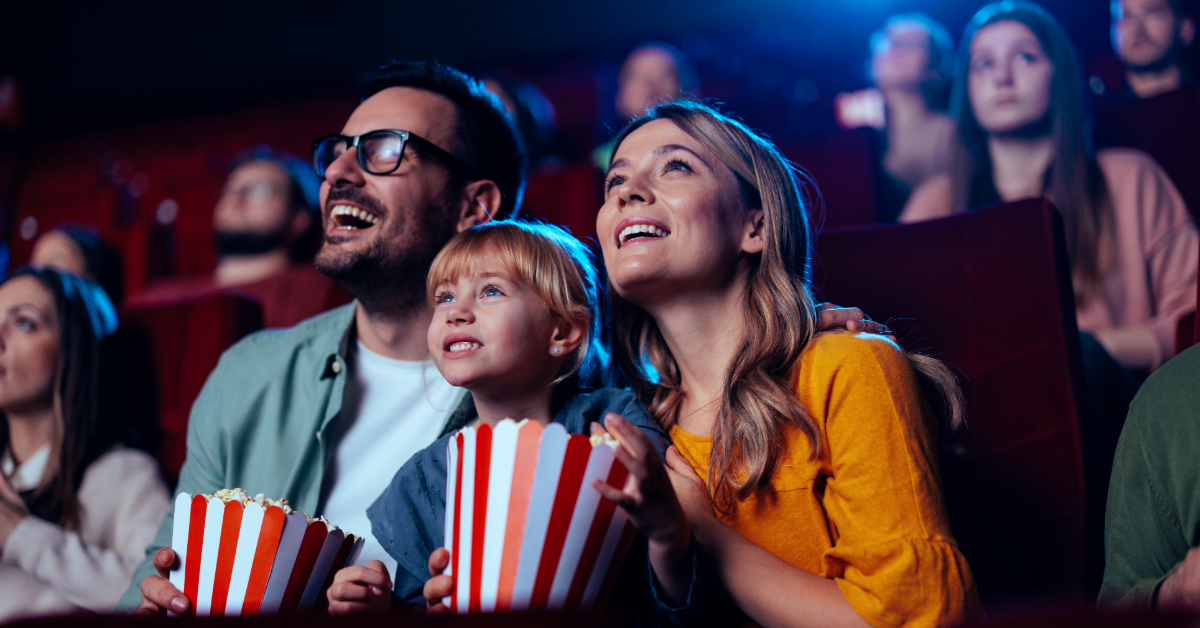 feat-img-julekino En familie sitter i en kinosal, smiler og holder stripete popcornbaljer, omgitt av andre publikummere.