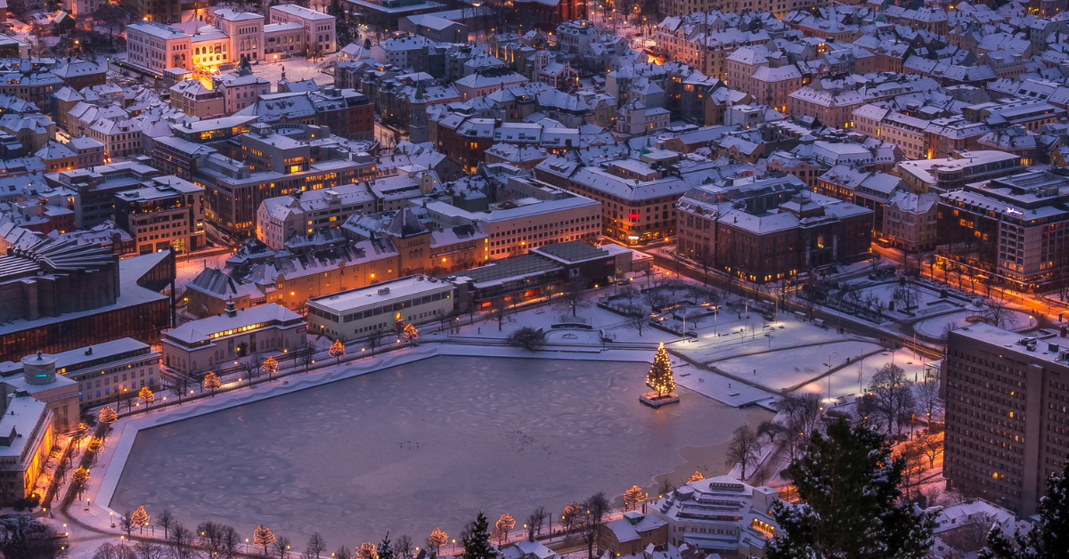 feat-img-mobilver Luftfoto av et snødekt bylandskap i skumringen, med en stor, frossen dam med et tent juletre i midten. Bygninger og gater er opplyst, og skaper en varm kontrast til snøen.