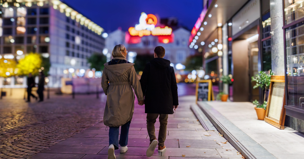 feat-img-opplev-sentrum Et par går hånd i hånd på en brosteinssti om natten, omgitt av opplyste butikker og uskarpt bylys.
