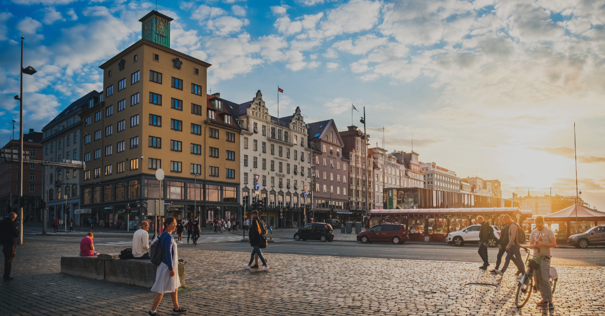 feat-img-paskeglede Folk som går og sitter på et travelt bytorg ved solnedgang, med historiske bygninger og en trikk i bakgrunnen under en delvis overskyet himmel.