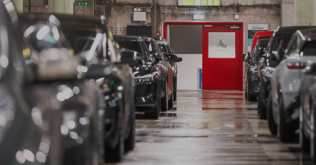 Biler parkert i et underjordisk parkeringshus med rød dør og nødutgangsskilt synlig i bakgrunnen.