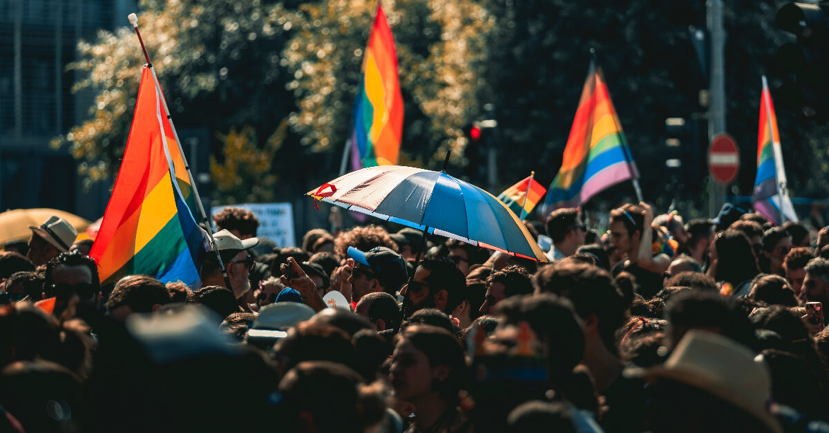 feat img template-1 (12) Det livlige publikummet på Bergen Pride fylte utendørsarrangementet, der flere pride-flagg vaiet ved siden av en person som holdt en blå og rød paraply.