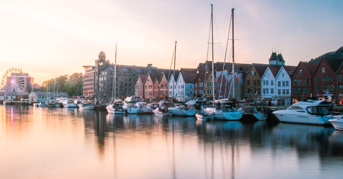 Båter som ligger til kai langs en vannkant med historiske trebygninger og et fjernt pariserhjul ved solnedgang skaper en pittoresk scene, forsterket av sommertilbud som gjør det til et uimotståelig reisemål.