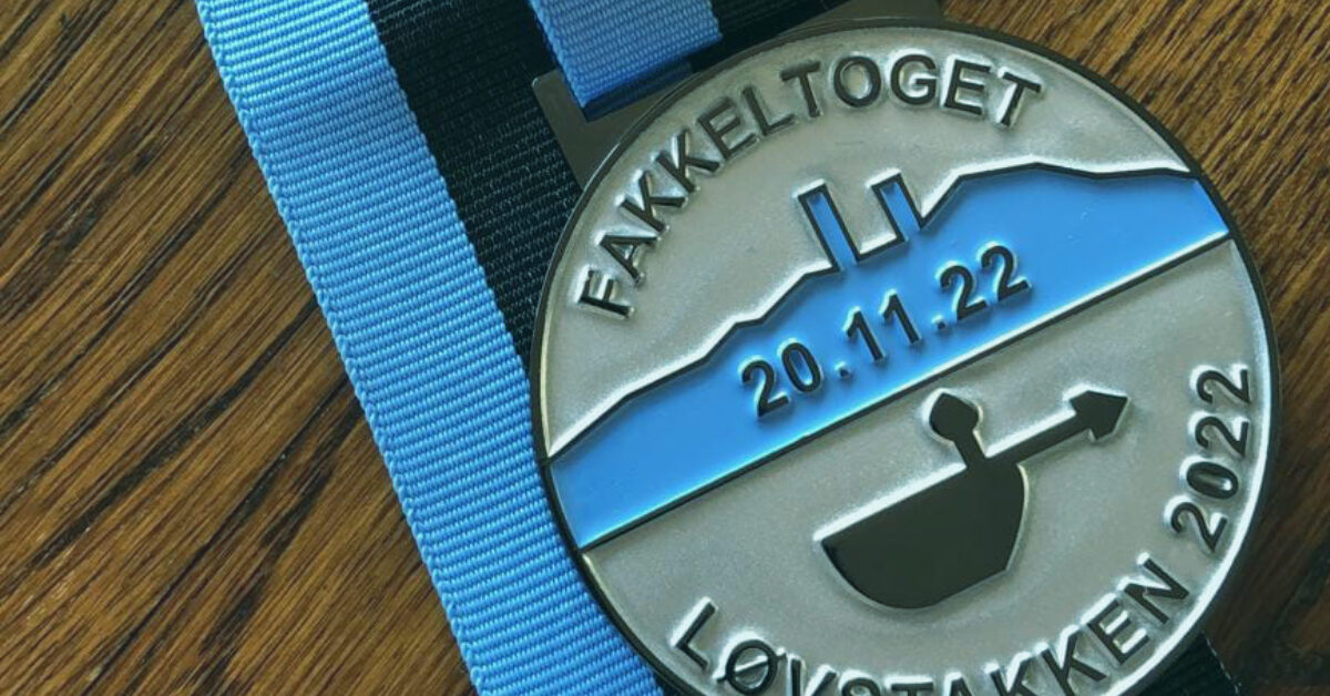 feat-img-template-1-3 En sølvmedalje med blått og svart bånd, påskrevet "FAKKELTOGET LØVSTAKKEN 2022" og datoen "20.11.22", med fjellsilhuett og flammende fakkeldesign.