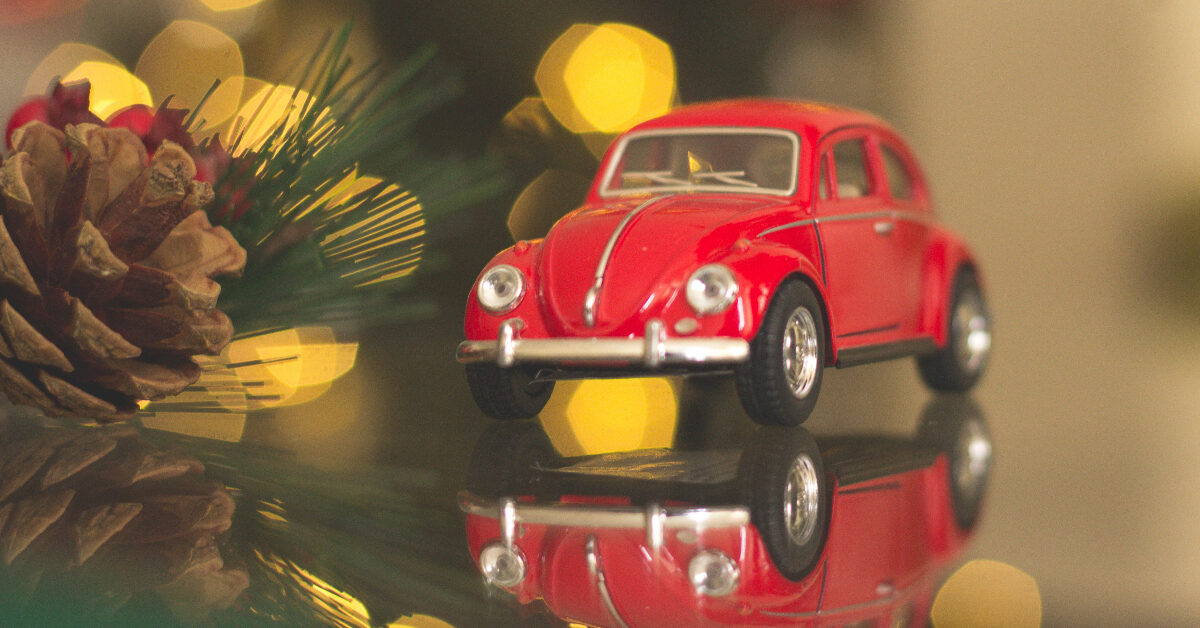 En rød lekebil som ligner en vintage Volkswagen Beetle reflekteres på en blank overflate, med en uskarp bakgrunn med kongler og gule lys, som fanger ånden av mobilitet og innovasjon som finnes i urbane garasjeprosjekter som Bygarasjen.