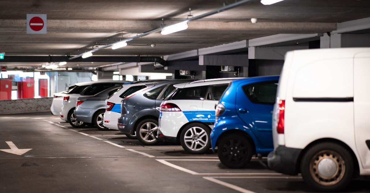 Biler og en varebil står parkert i et svakt opplyst innendørs parkeringshus, der bergens parkeringsskilt lyser under taklysene.