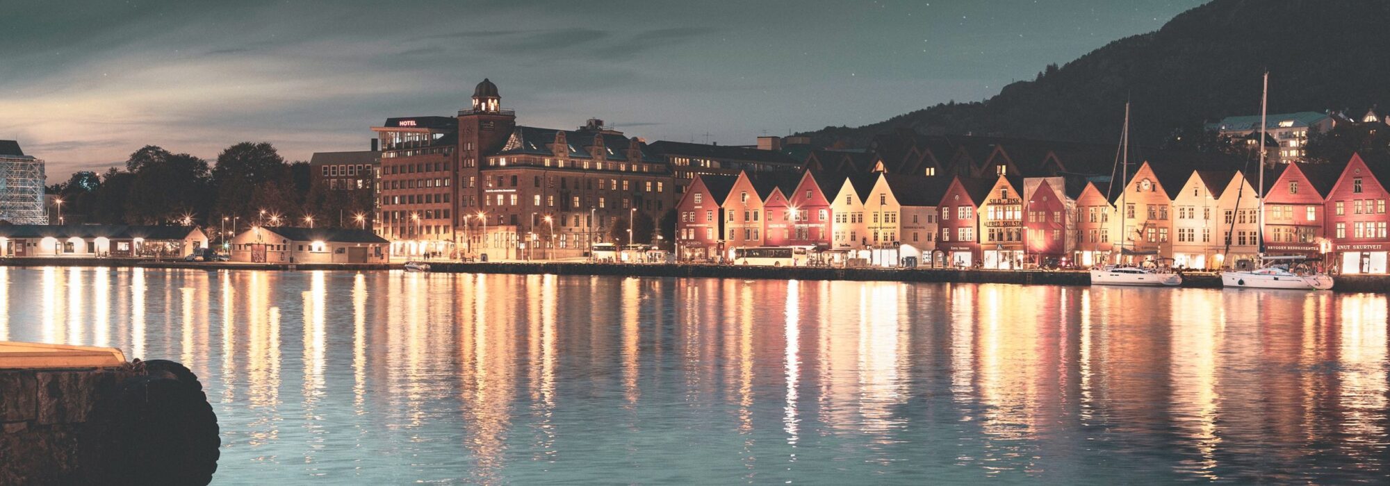 momentista-reR1qazgsBM-unsplash En rad med fargerike bygninger langs en vannkant under en skumringshimmel, med refleksjoner som glitrer på det rolige vannet. Åsene reiser seg i bakgrunnen under en stjernehimmel.