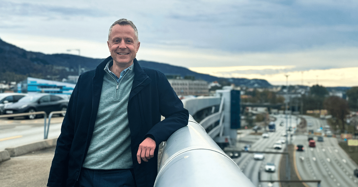 ny daglig leder feat img En mann i jakke og genser står smilende med et bylandskap og en motorvei synlig i bakgrunnen under en overskyet himmel.