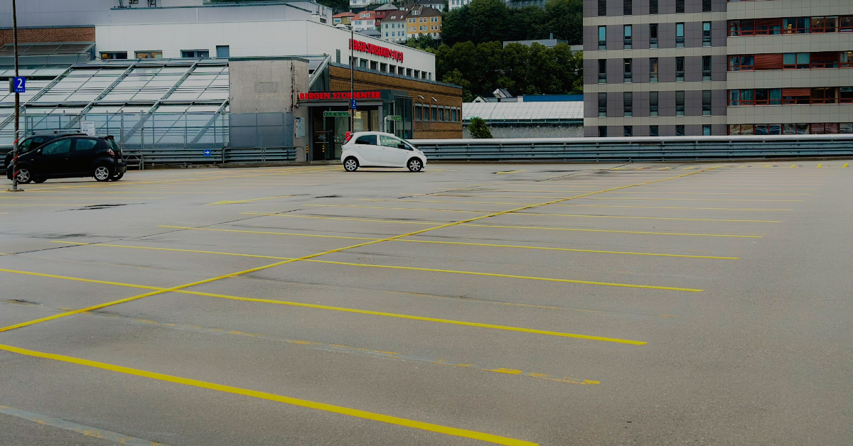 På en nesten tom parkeringsplass på taket sitter to biler – en svart og en hvit – stille og tilbyr rikelig med bedre plass. I bakgrunnen markerer bybildet i Bergen og en ås utsikten, og gjenspeiler bekvemmeligheten til nærliggende parkeringsmuligheter som ByGarasjen.