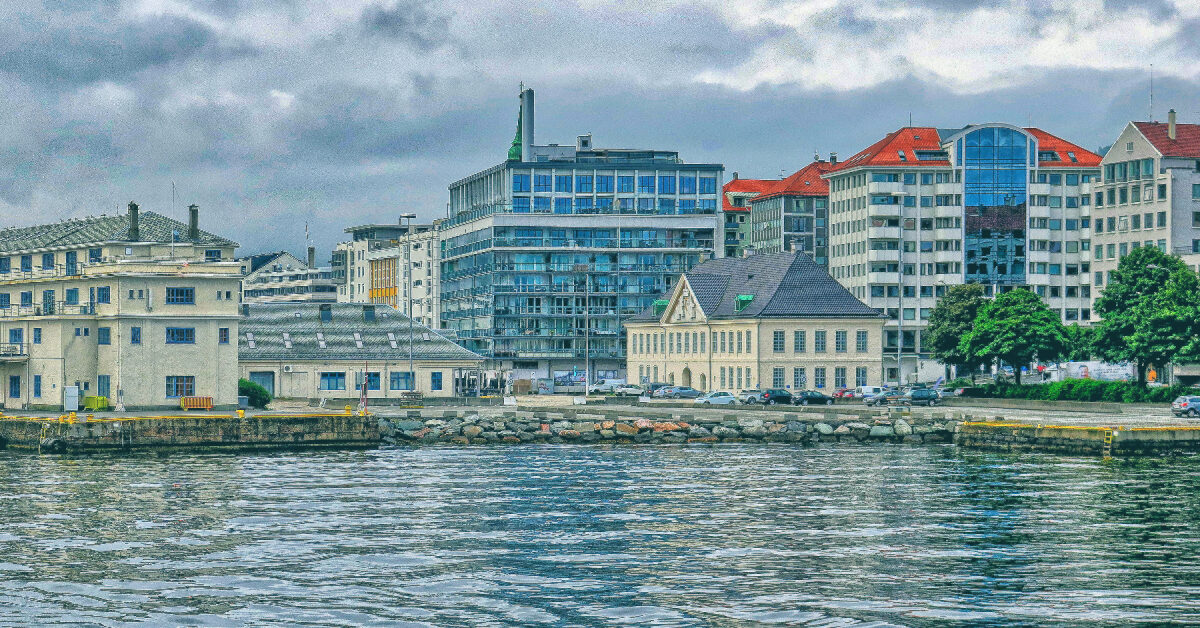 tollbodkaien Kystbybildet har en blanding av moderne og historiske bygninger under overskyet himmel, med vann i forgrunnen og Tollbodkaien som fører til de sjarmerende bryggene.