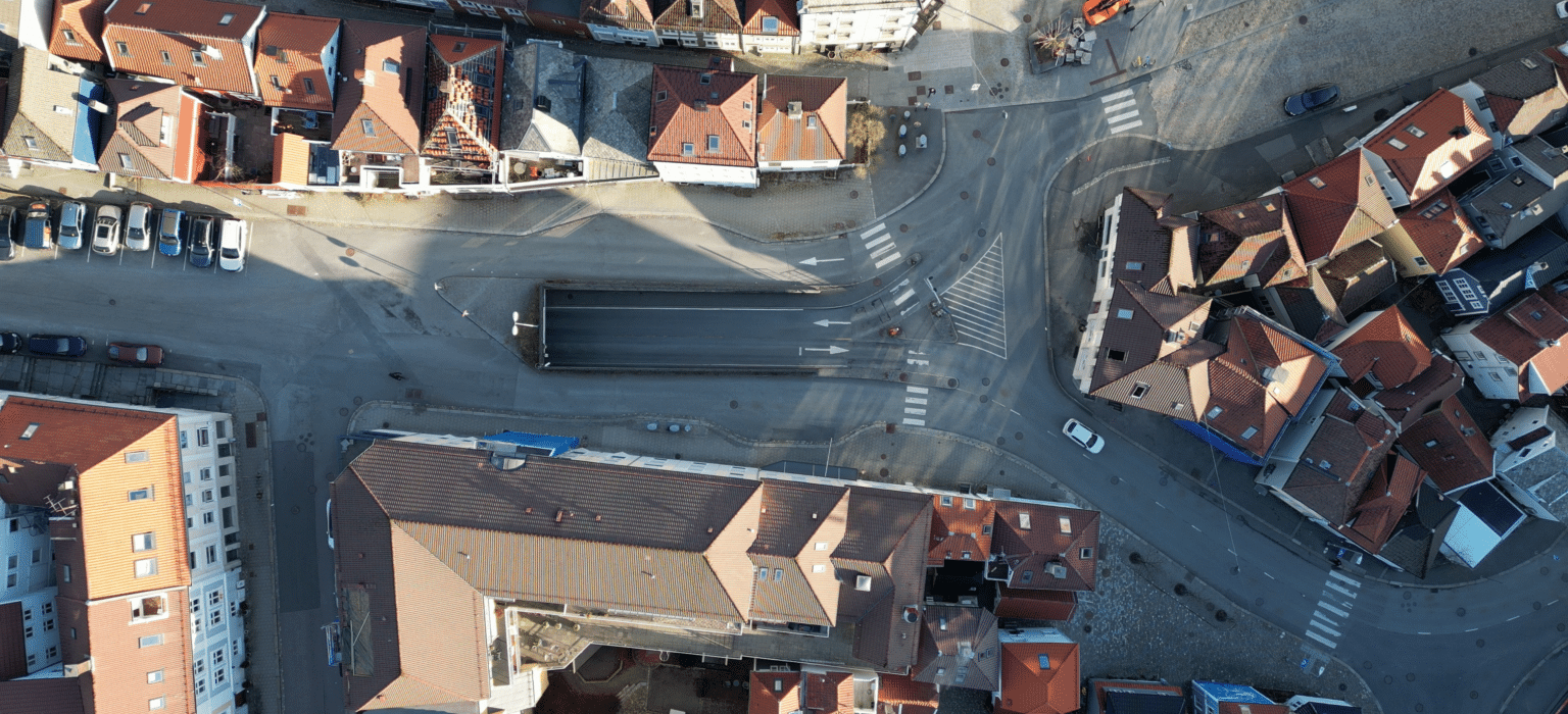 Luftfoto av et lite byområde med bygninger med røde tak, kryssende veier og en overbygd gangvei.