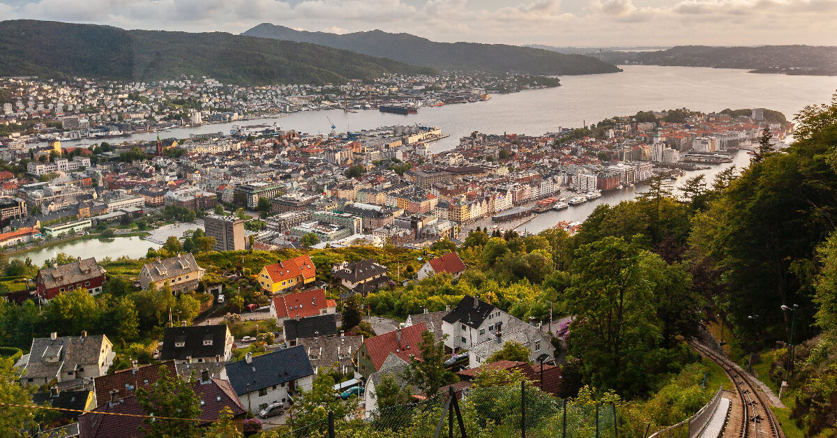Luftfoto av Bergen sentrum, en kystby med bygninger, en havn og omkringliggende åser under overskyet himmel. Et jernbanespor går gjennom forgrunnen, med grøntområder og hus i nærheten. Parkeringsfasiliteter er synlige midt i det travle bylandskapet.