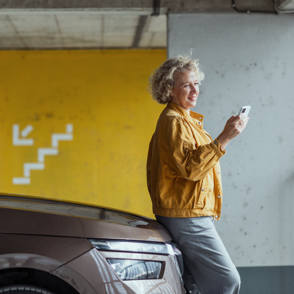 Bilde av en kvinne som lener seg inntil en bil i et parkeringshus, mens hun kikker på mobilen sin og smiler