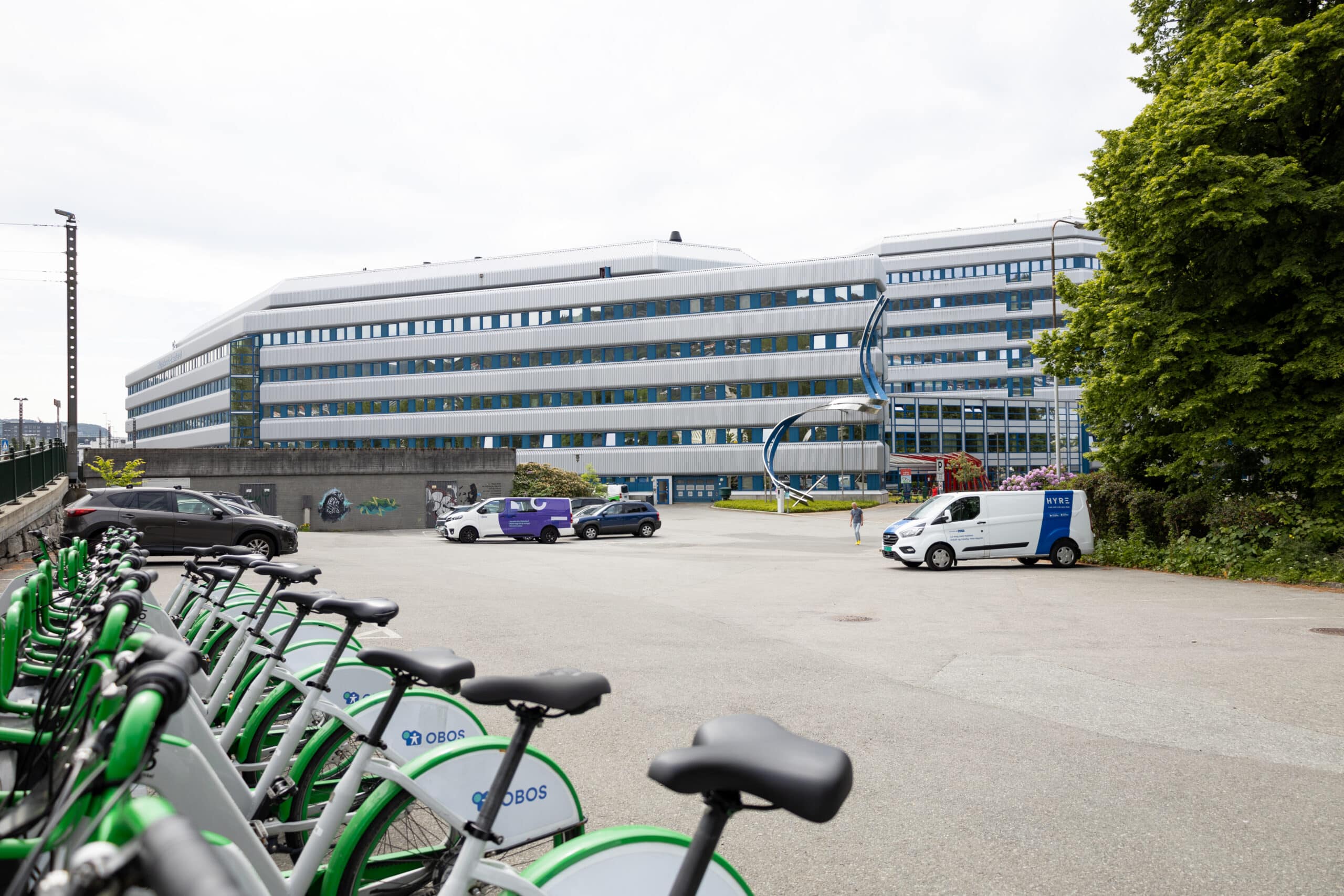Rekke med parkerte sykler foran et stort moderne kontorbygg, med biler synlige på parkeringsplassen og en skulptur nær inngangen. Trær er til høyre.