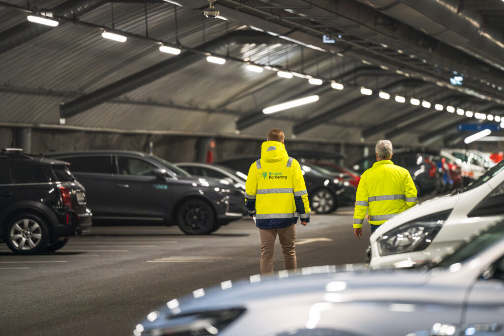 Bilde av to menn sett bakfra som går i KlosterGarasjen. De har på seg synlighetstøy med logoen til Bergen Parkering på ryggen.