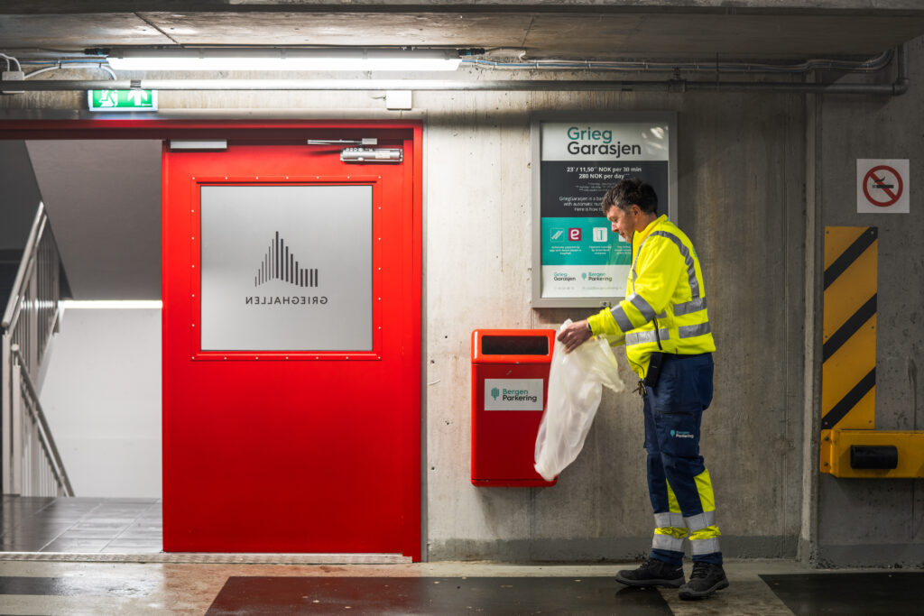 Bilde av en mann som tømmer søppelspann i GriegGarasjen. Han har på seg synlighetstøy med logoen til Bergen Parkering på ryggen.