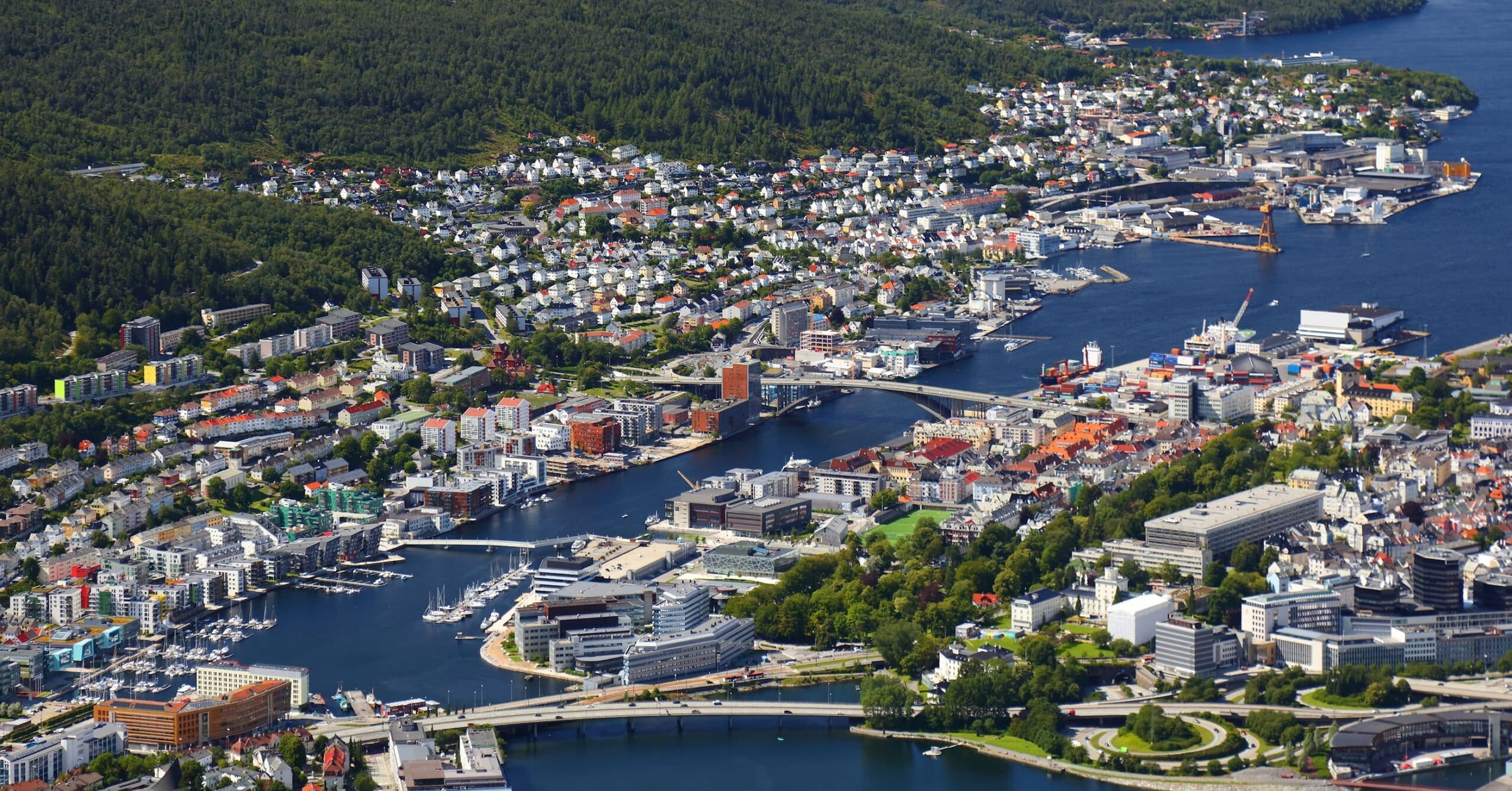 Luftfoto av en kystby med tettbebyggelse, en havn, broer og skogkledde åser i omgivelsene.