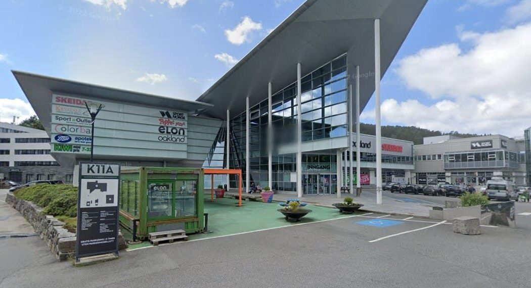Moderne næringsbygg med skråtak, store glassvinduer og diverse butikkskilt. Flere biler er parkert på plassen. Grønt sykkelområde i forgrunnen. Overskyet himmel.