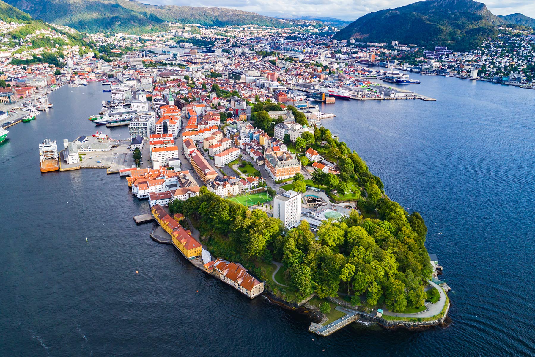 Luftfoto av Bergen, Norge, som viser et kystbybilde med fargerike bygninger, frodige grøntområder og omkringliggende vann, plassert mellom fjellterreng.