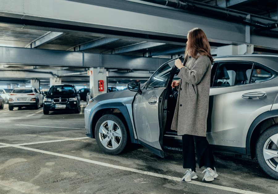 feat-img-BG-plan-1-sor En person iført lang frakk står ved siden av en åpen bildør i et parkeringshus med flere parkerte biler.
