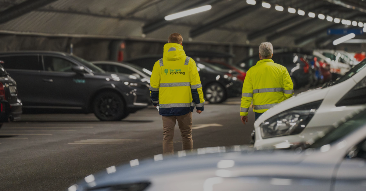 Bilde av to menn sett bakfra som går i KlosterGarasjen. De har på seg synlighetstøy med logoen til Bergen Parkering på ryggen.