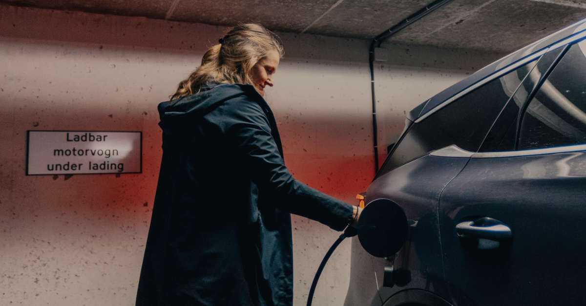 Voksen kvinne setter ladekabel inn i en bil i et parkeringshus