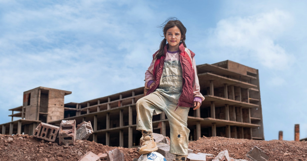 Bilde av et jentebarn rundt 8 eller 10 år som står foran et ruinert bygg. Bildet ser ut til å være tatt i et krigshjertet land.