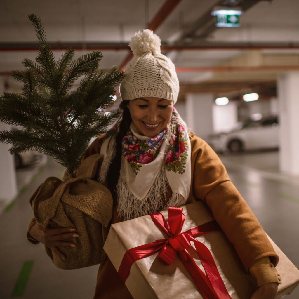 Bilde av en kvinne i et parkeringshus. Hun har mange julegaver i hendende, lue på hodey, og smiler mens hun går bortover.
