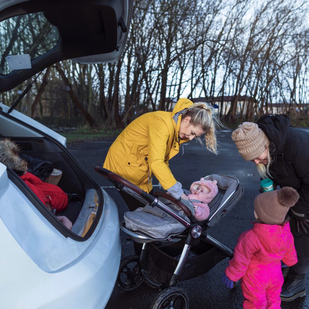 Bilde av to kvinner med to små barn, det ene i barnevogn. Alle står ved baksiden på en bil med bagasjerommet åpent.