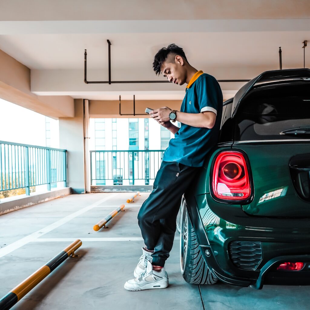 Bilde av en ung mann som lener seg inntil en parkert bil i et parkeringshus. Han holder en smarttelefon i hendene og ser ned mot den.