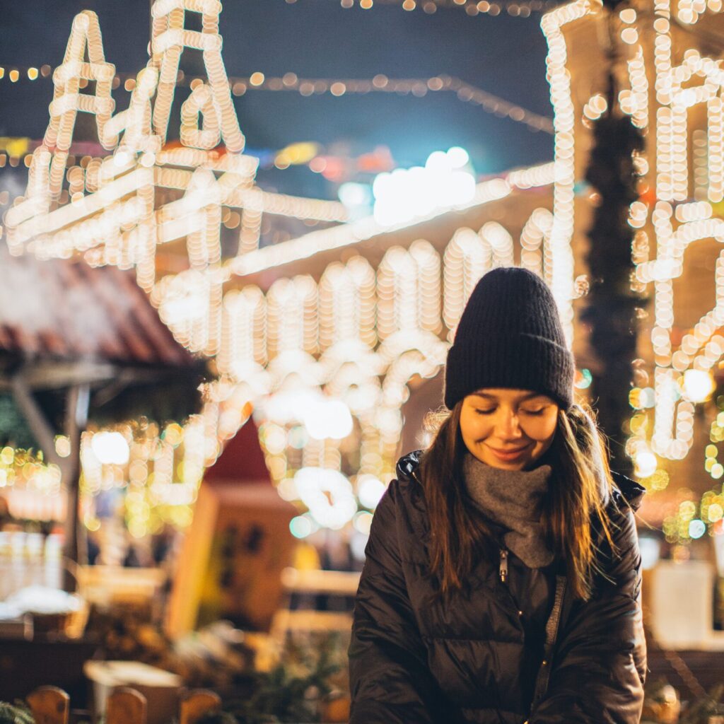Bilde av en dame ute på et julemarked, med masse boder og julelys i bakgrunnen.