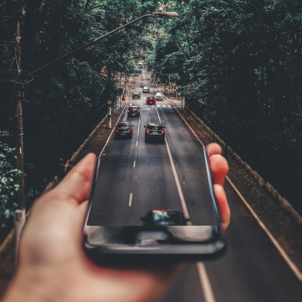 Bilde av en hånd som holder en smarttelefon i forgrunnen. Bildet manipuleres til å bli en bilvei i landlige omgivelser.