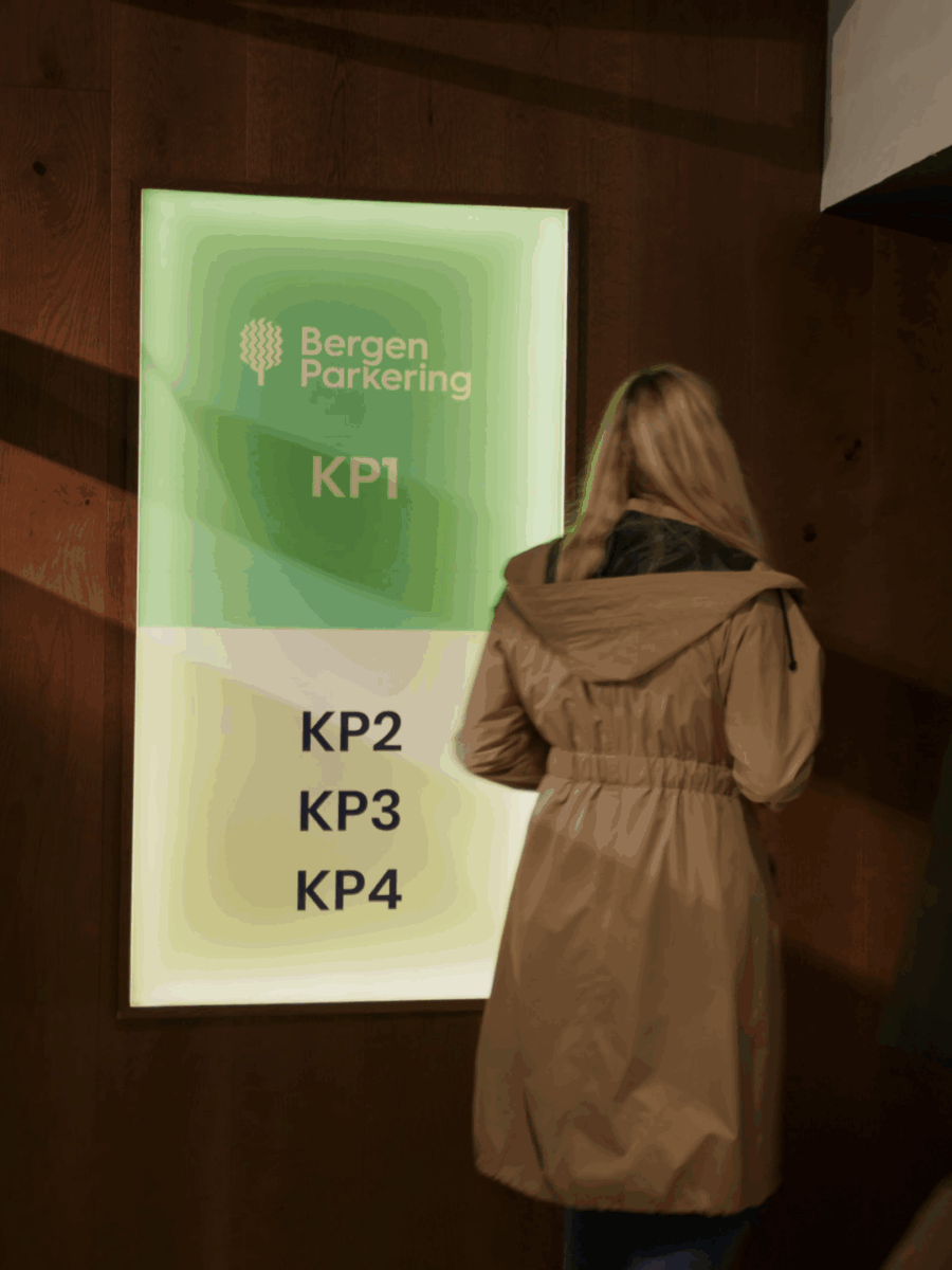 En kvinne i brun frakk går mot et opplyst Bergen Parkering-skilt på Bergen Kino.
