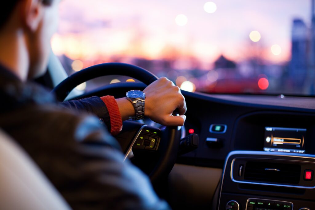 Bilde av en mann i førersetet i en bil. Bildet er tatt fra baksetet. Han har ene hånden på rattet og i bakgrunnen ser man en trafikkert vei i solnedgang.
